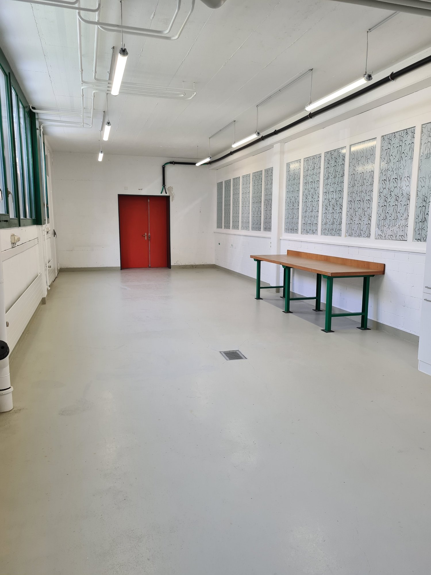 Wide empty room with white walls, white floor, red door, a wooden table, white pipes