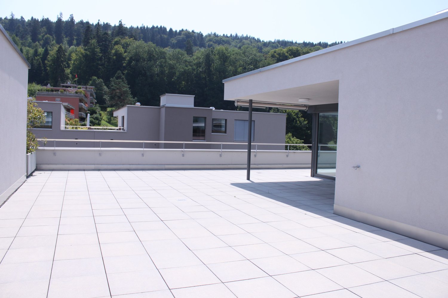 Terrace with tile flooring, surrounded by buildings, overlooking a forested hill, with a balcony on the left side