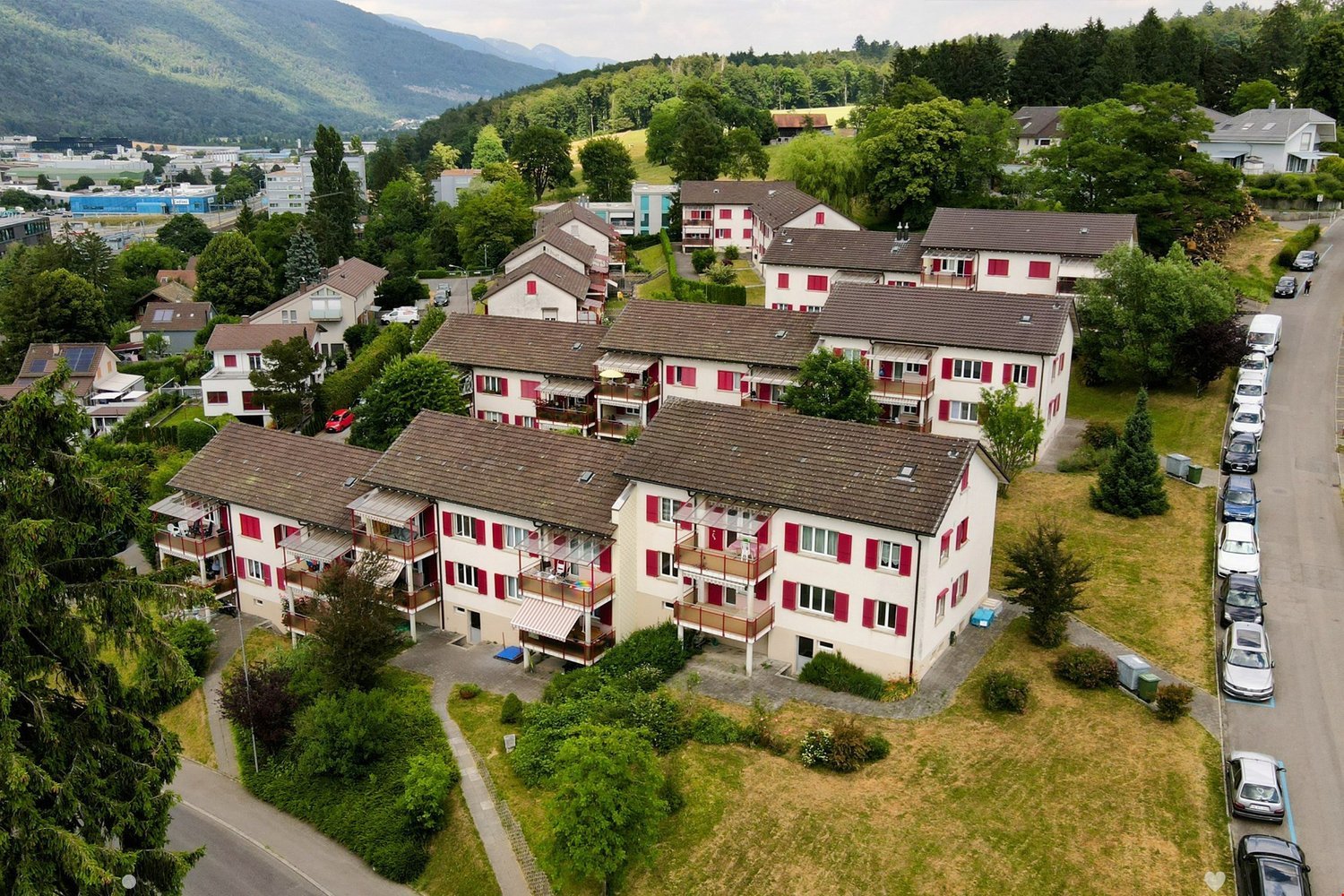 Apartment buildings with multiple floors, balconies, brown roofs, parking spaces, greenery, surrounding mountains