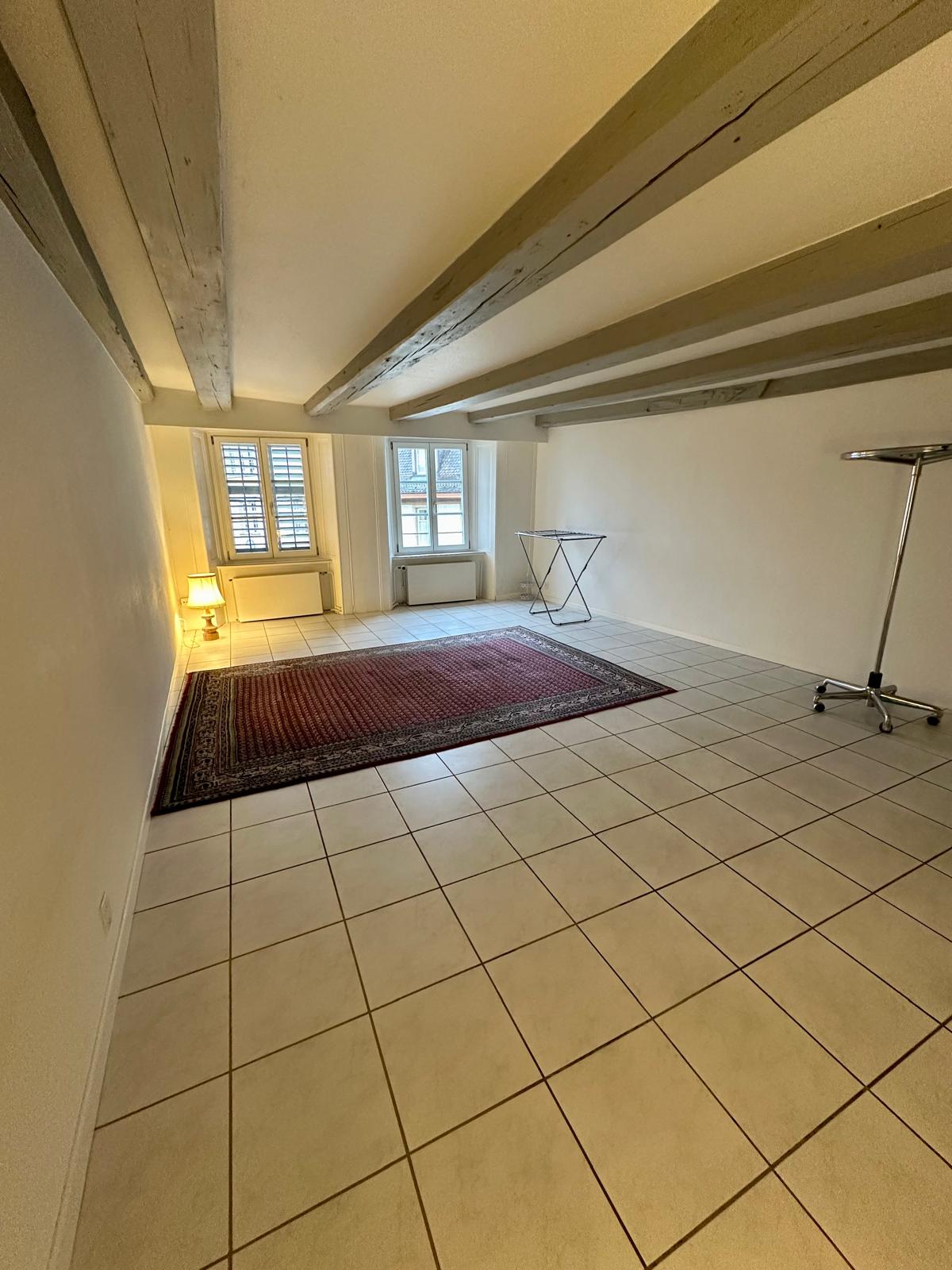 Empty room with tiled floor, two windows, ceiling beams, a rug, and a few small tables