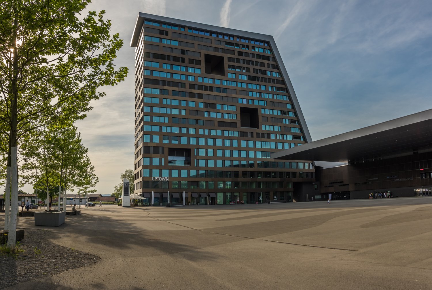 Modern high-rise building with glass facades, large triangular design, multiple entrances, surrounded by trees and concrete parking lot, signage reading UPTOWN