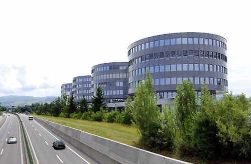 Multi-story office building with curved glass facade, surrounded by trees and a highway with cars passing by
