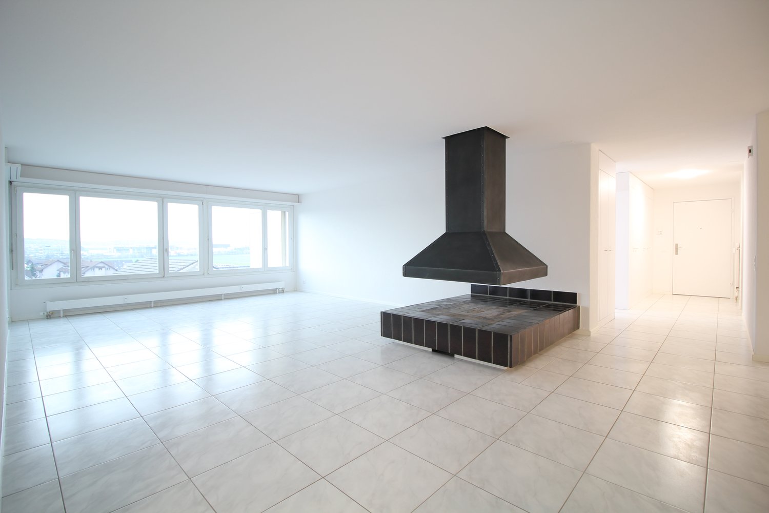 large empty room, white tile flooring, tall windows, fireplace in the center, metal chimney