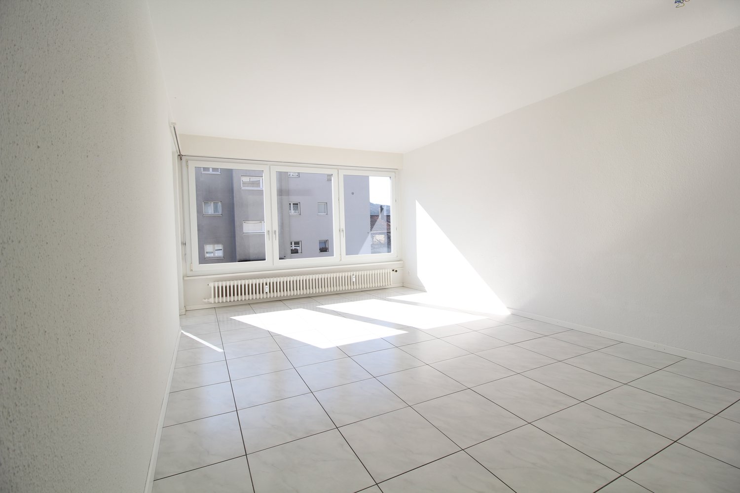 Empty room with white walls, white tiled floor, large window, white radiator