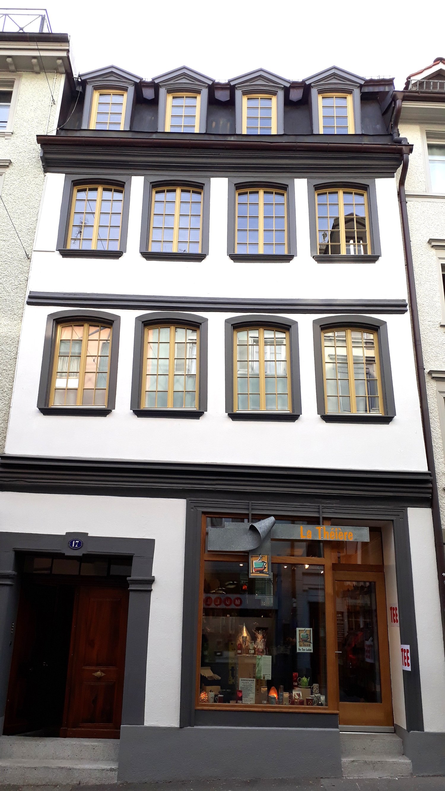 2-story building, white walls, yellow window frames, black roof, shop with glass windows, wooden door