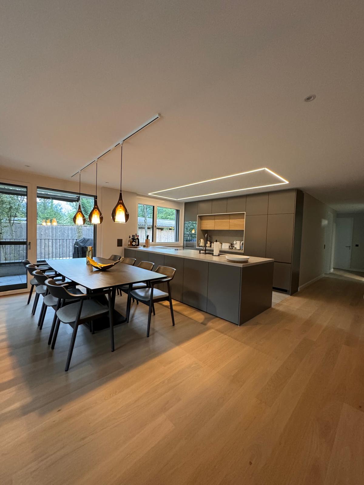 The image shows a modern, open-concept kitchen and dining room. The kitchen features dark gray cabinets and a large island with a built-in dishwasher. The dining area has a long wooden table with black chairs. Pendant lights hang above the dining table, a
