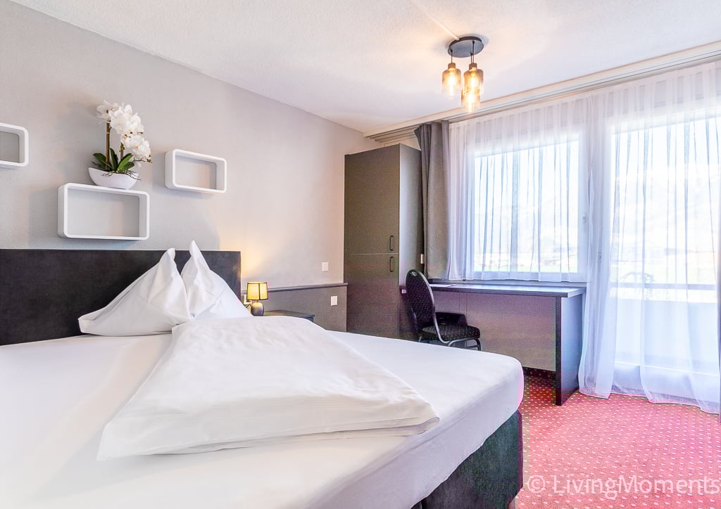 Spacious bedroom with a large bed, white bedding, and a desk area. The room has large windows with curtains, providing natural light. There are shelves on the wall and a decorative plant. The room has a modern and cozy feel.