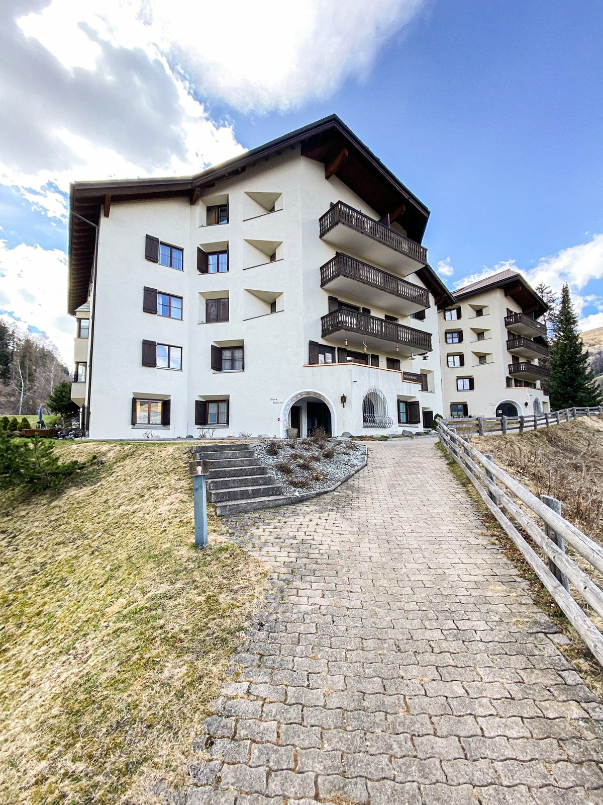 A multi-story apartment building with a white exterior, balconies, and a stone pathway leading up to the entrance. The building is surrounded by trees and has a scenic mountain backdrop.