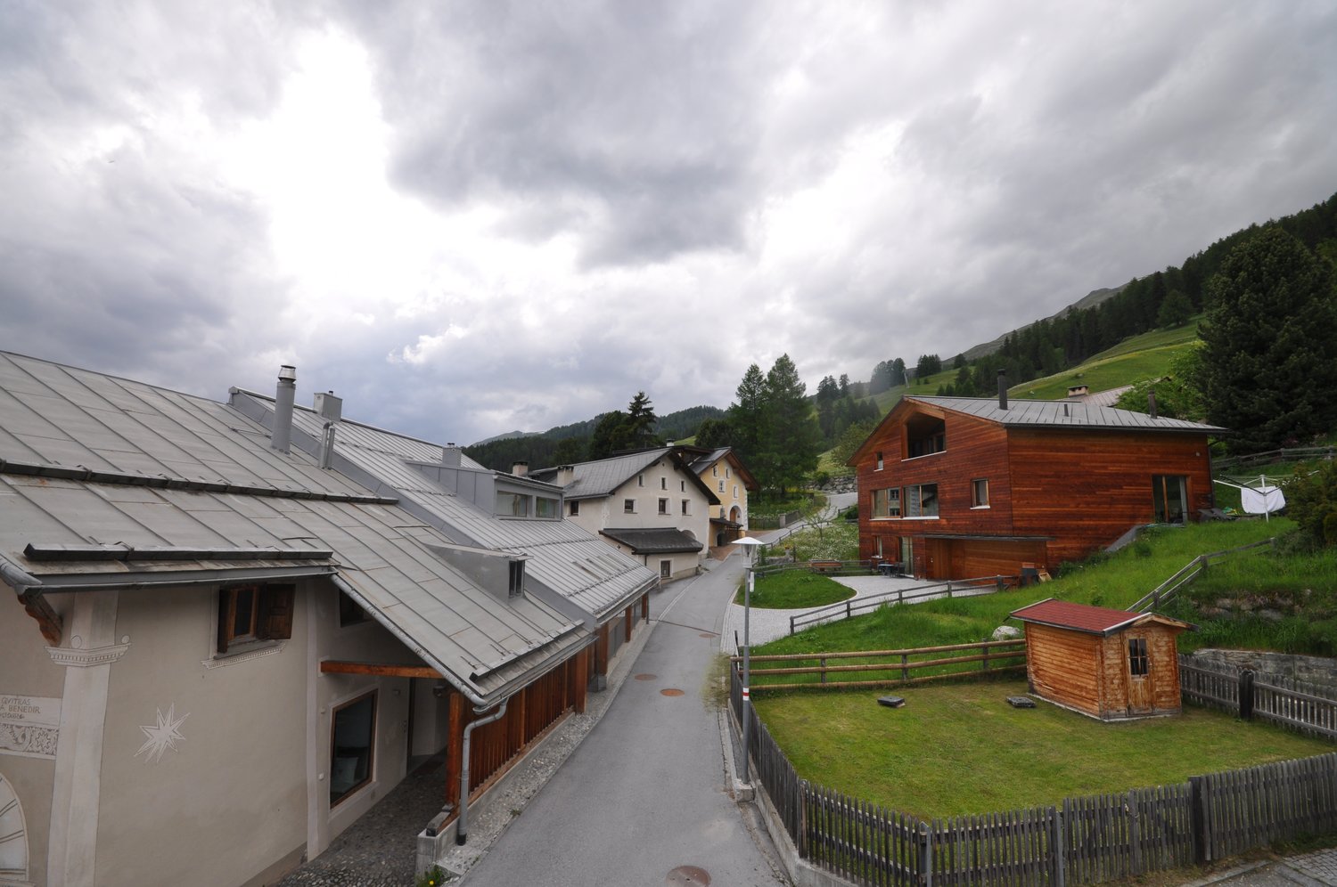 The image shows a picturesque village nestled in a mountainous landscape. The buildings have a traditional alpine architecture with wooden facades and metal roofs. There are several houses, some with balconies, and a path running through the village. The