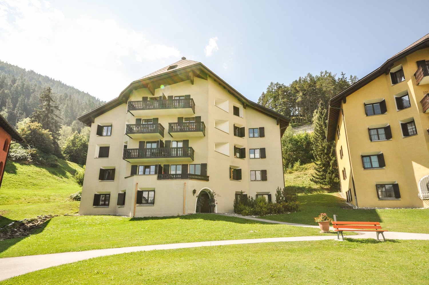 4-story apartment building with yellow exterior, balconies, surrounded by green grass and trees, located in a mountainous, forested area