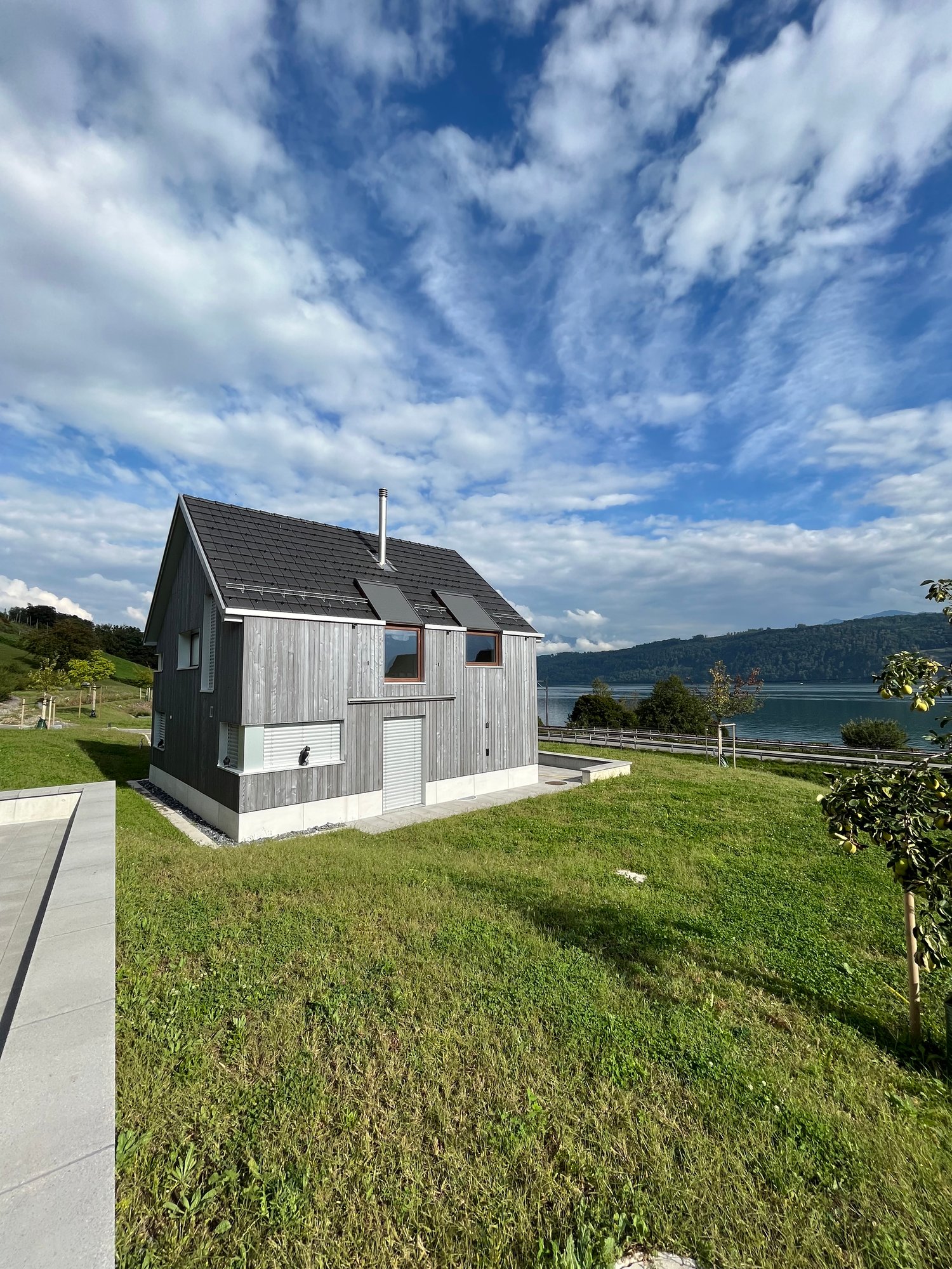 A modern, two-story wooden house with a slanted roof and a chimney, situated on a grassy hill overlooking a body of water and surrounded by mountains and cloudy blue sky.