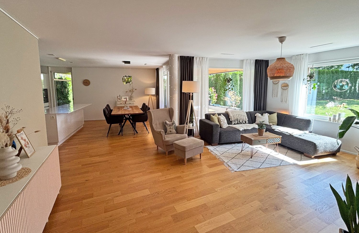 Wooden floor, modern grey sofa, black dining table, floor lamp, white cabinets, potted plants, rug
