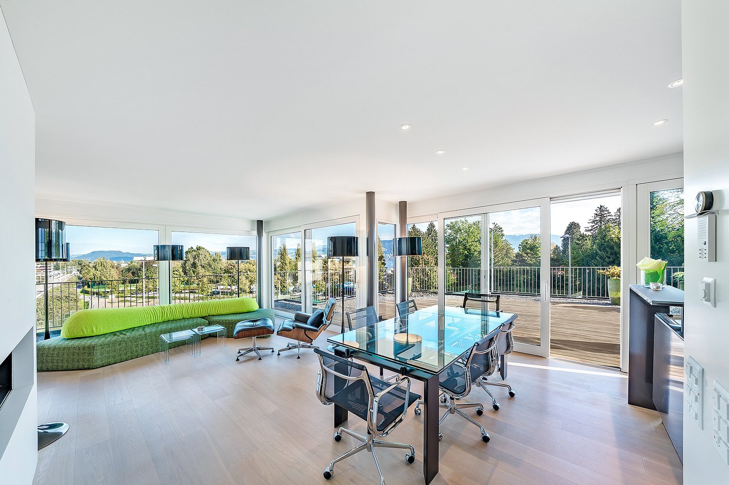 large windows, balcony with railings, green couch, glass table, office chairs, lamps, hardwood floor