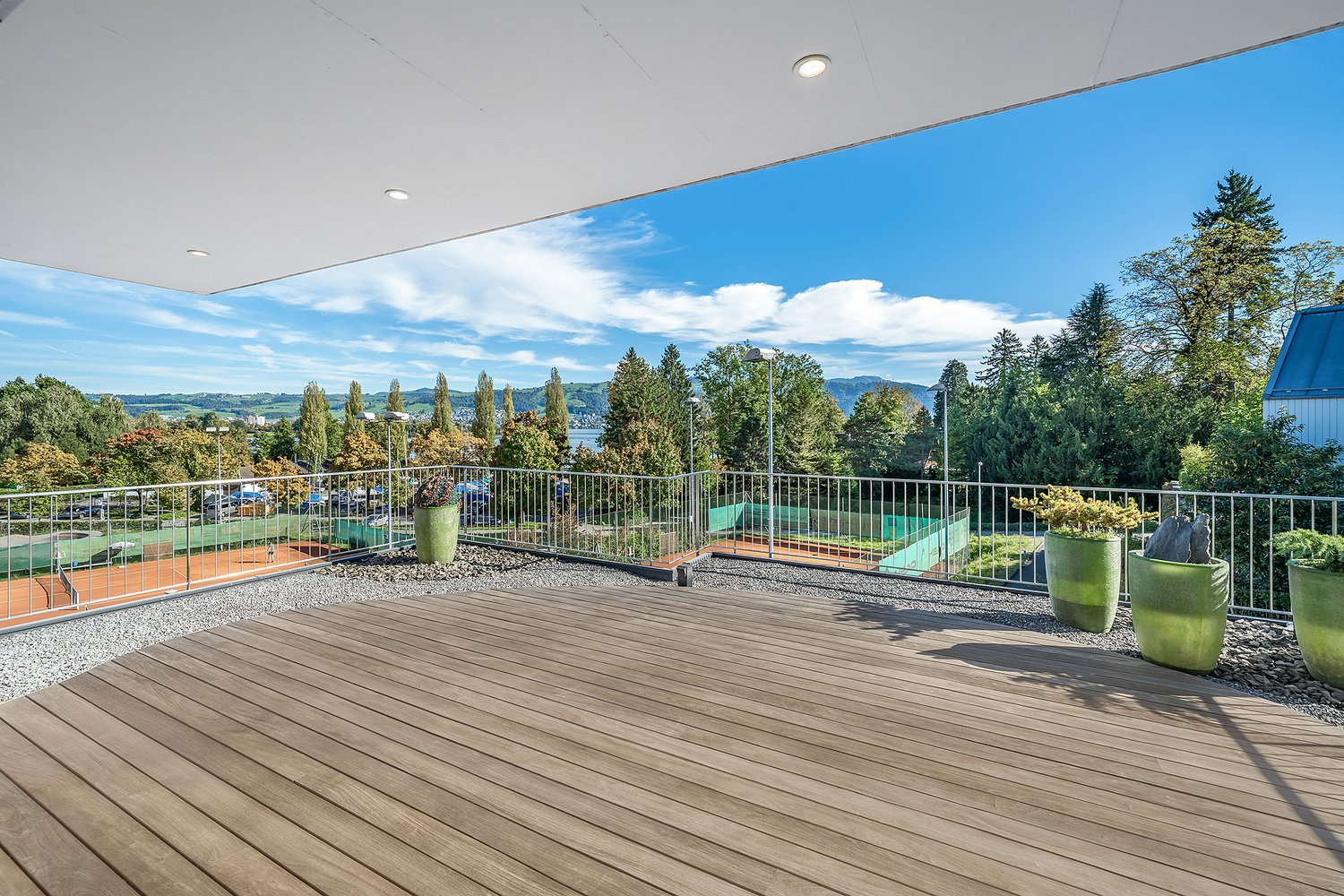 Balcony with metal railings, wooden floor, gravel path, 3 green potted plants, panoramic view of landscape