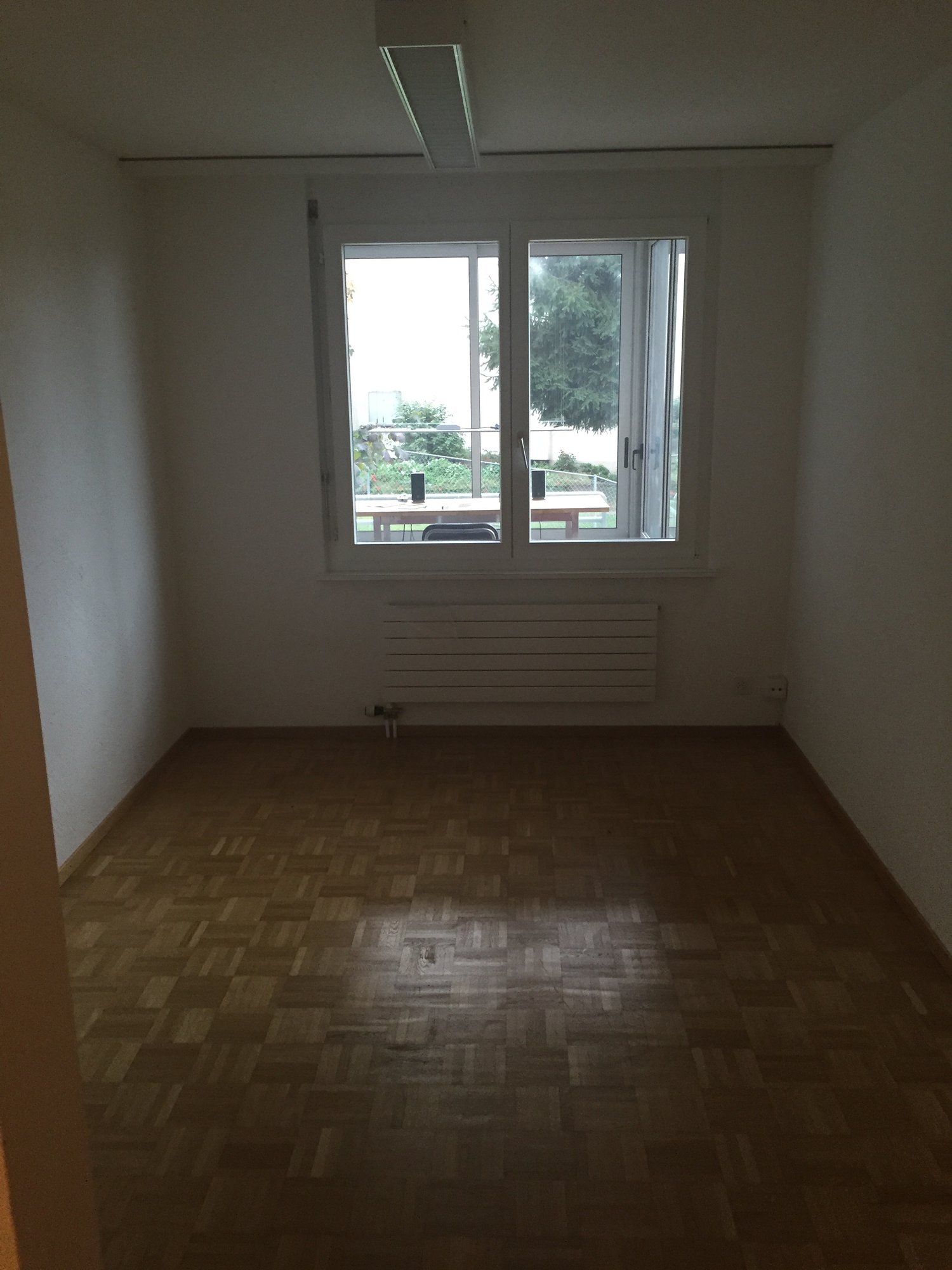 This image shows a room with large windows overlooking a balcony or terrace. The room has white walls and a patterned tile floor. There is a radiator installed along the wall below the windows.