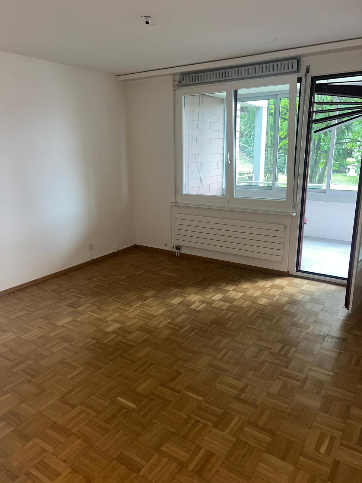 Spacious room with large windows, hardwood parquet flooring, and a balcony or terrace visible through the windows.