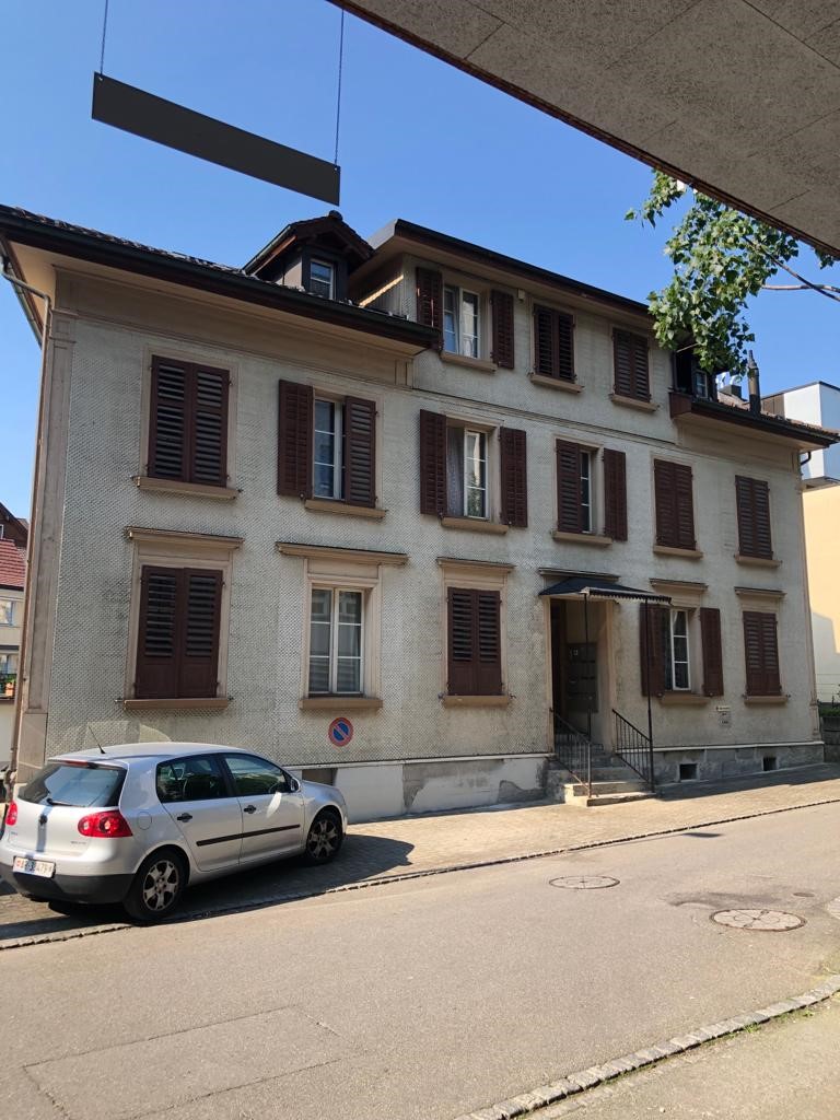 A multi-story residential building with a traditional European architectural style. The building has a stucco exterior, brown shutters on the windows, and a sloped roof. There is a car parked on the street in front of the building.