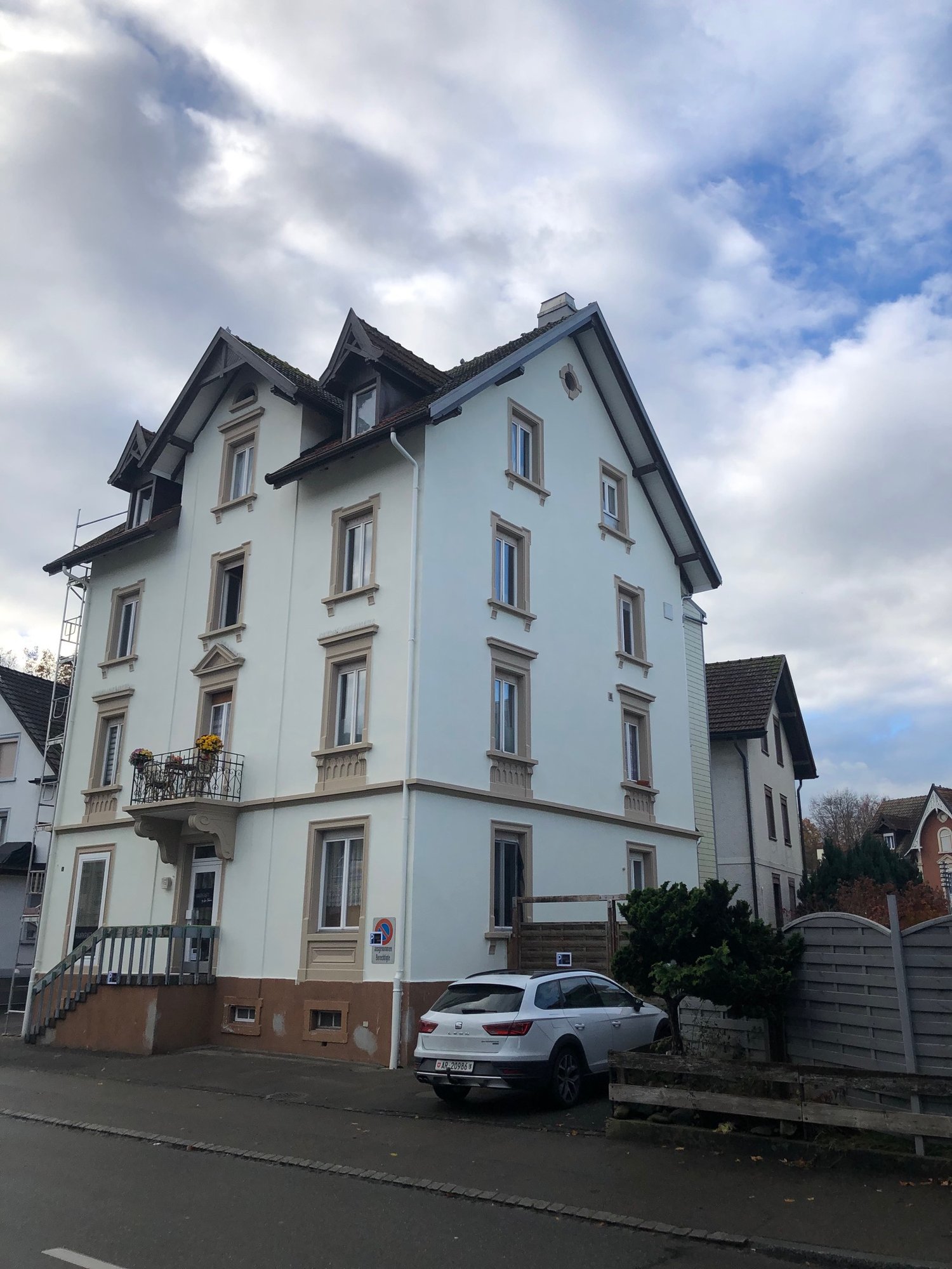 large multistory white building, brown trim, multiple windows, stairs leading to the entrance, balcony on the second floor, a car parked in front