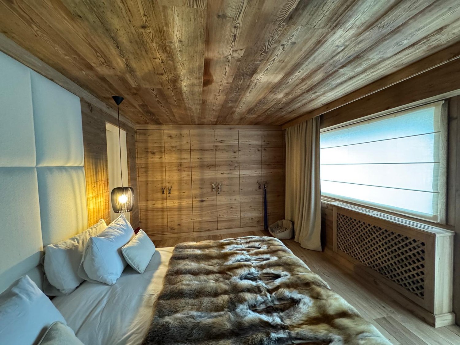 Rustic wooden walls and ceiling, large windows with a view, fur blanket on the bed, bedside lamps, curtains