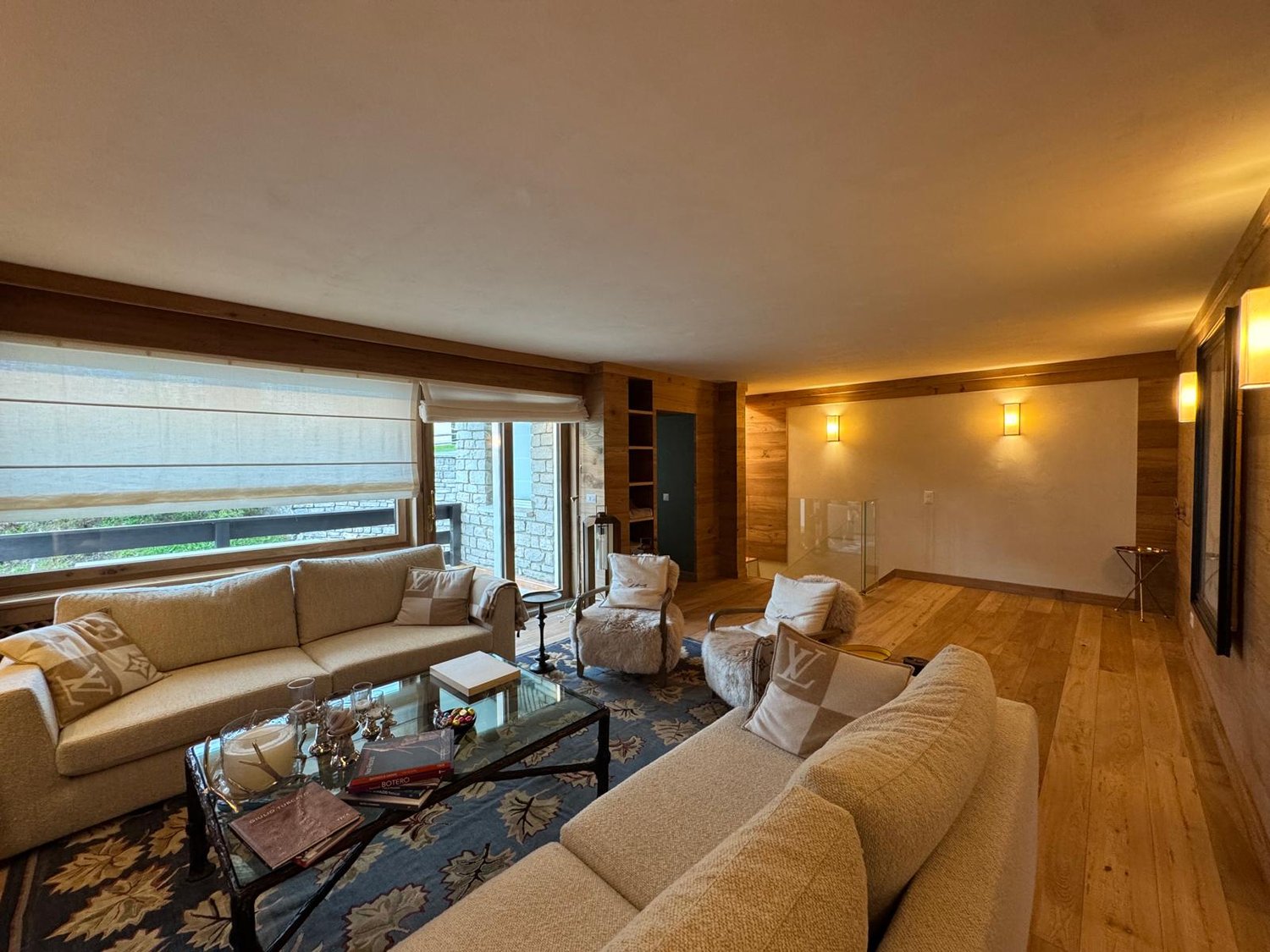 Spacious living room with large windows, hardwood floors, plush beige sofas, coffee table, and a fireplace. The room has a warm, cozy atmosphere with wooden accents and decorative elements.