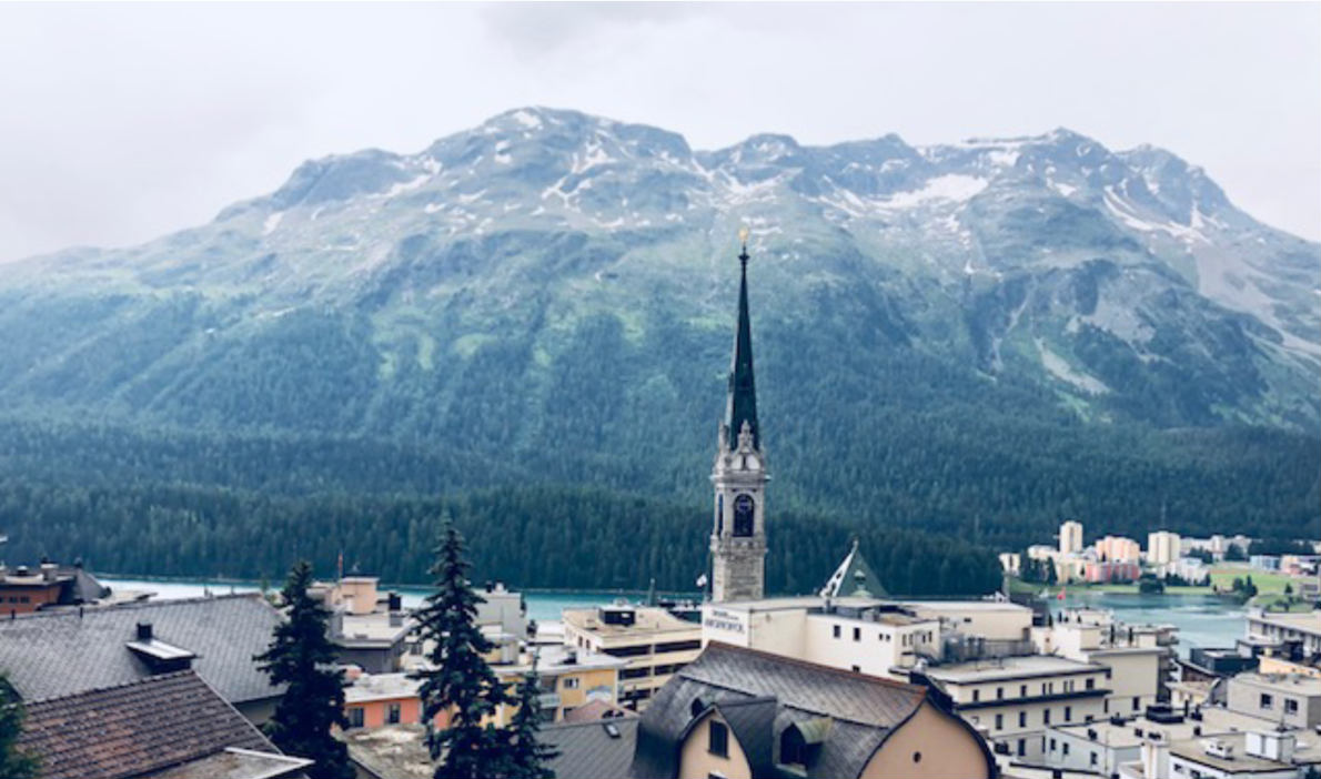 The image shows a scenic view of a mountainous landscape with a church steeple in the foreground. The town appears to be nestled among the snow-capped peaks, with a mix of residential and commercial buildings visible. The overall scene has a picturesque,