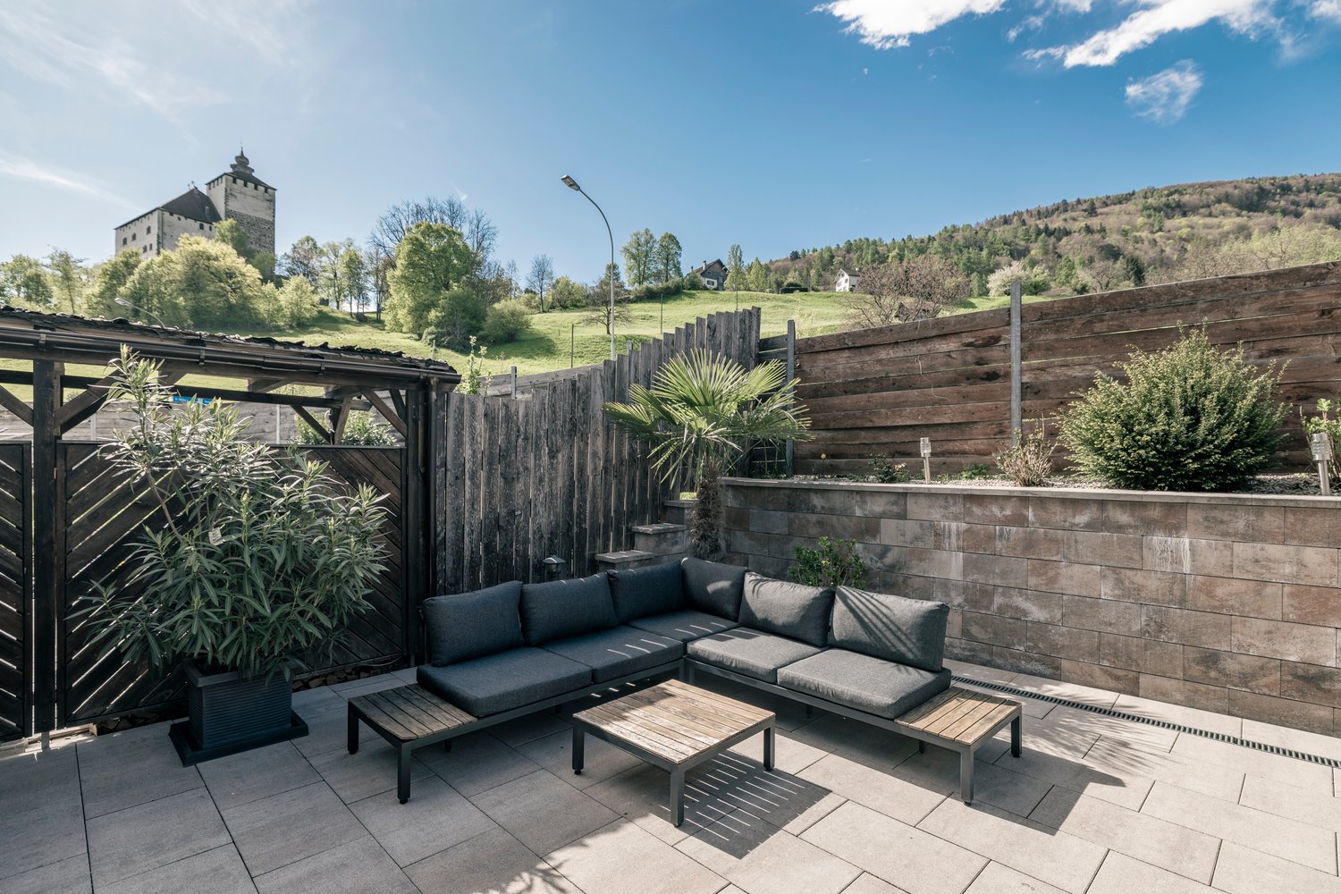 Outdoor patio with wooden deck, black patio furniture including a sectional sofa and coffee table, surrounded by a wooden fence and lush greenery. In the background, there is a castle tower visible on a hill.