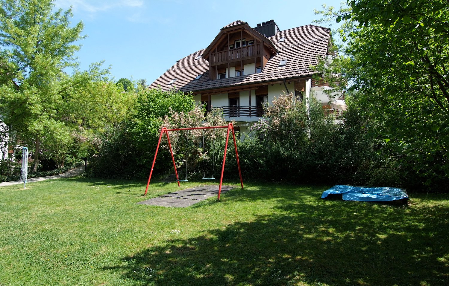 House with brown roof and balconies, surrounded by greenery. A playground with a swing and a blue tarp on the ground.