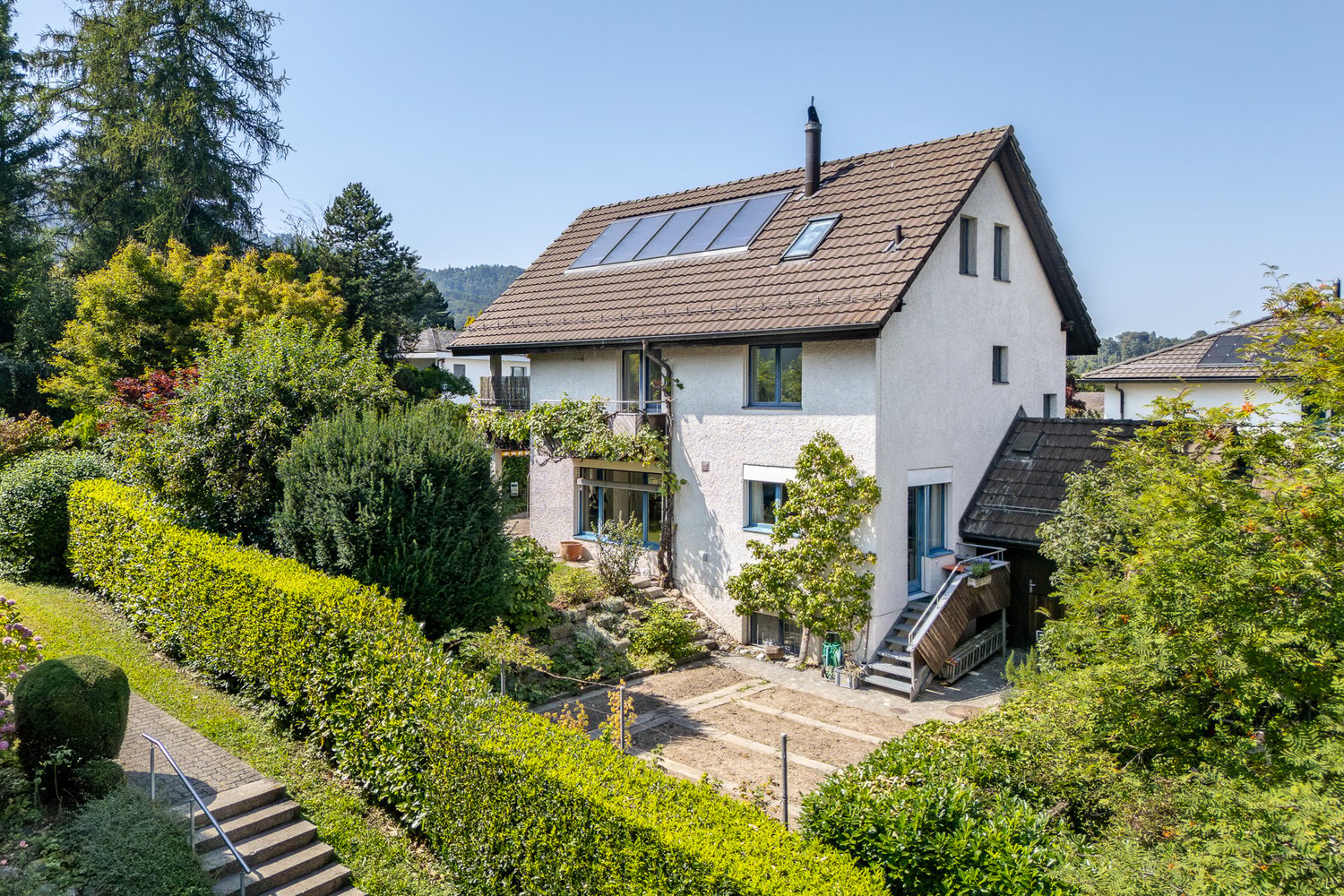 2 story house, brown roof, solar panels, white walls, surrounded by trees and shrubs, balcony