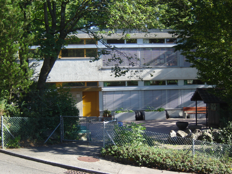 Modern house with yellow door, white walls, multiple windows, surrounded by trees and plants, small garden with bench and plants