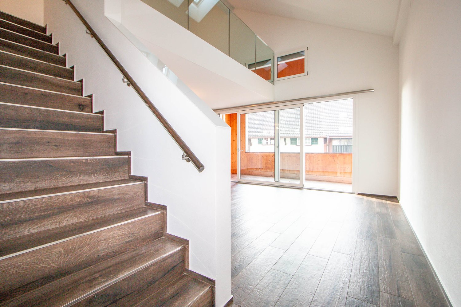 Spacious open-plan living area with wooden staircase, large windows, and sliding glass doors leading to a balcony or terrace.