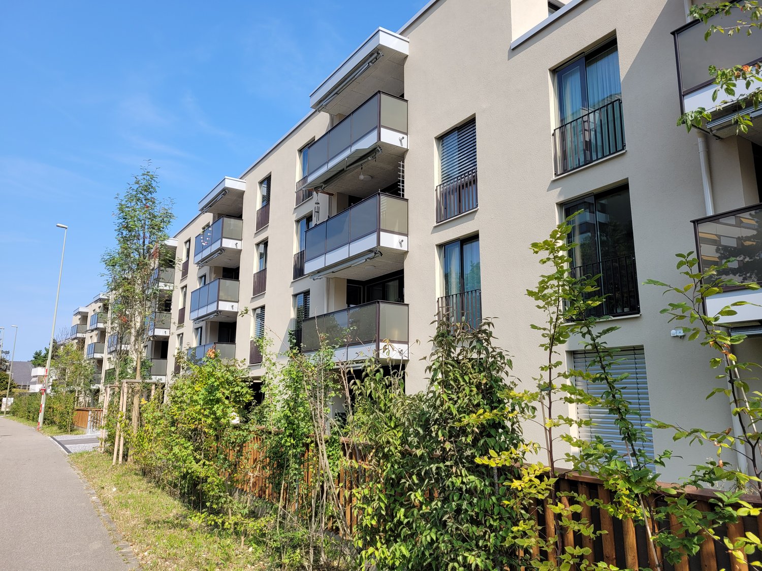 A multistory apartment building with balconies, beige facade, and wooden fence. There are shrubs and trees in front. A streetlight is on the left side.