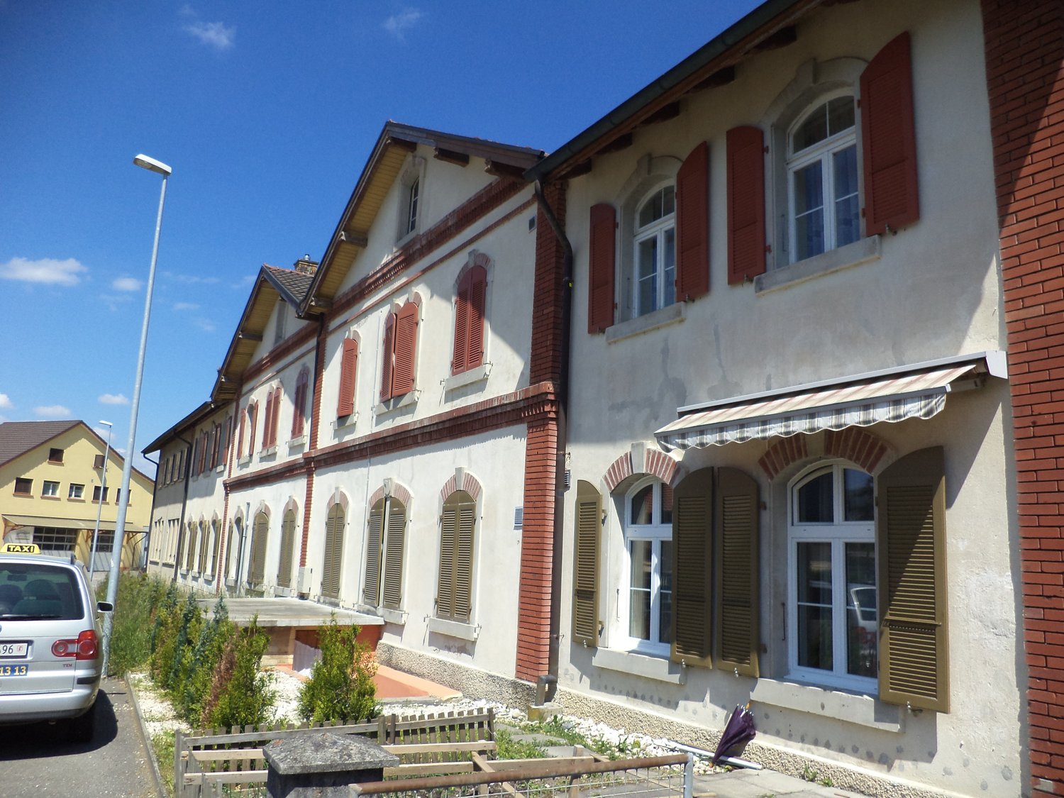 A multi-story building with white walls, red bricks, and multiple windows, some with shutters. It has a gray concrete awning over one of the windows, a gray metal fence and a car parked on the street in front.