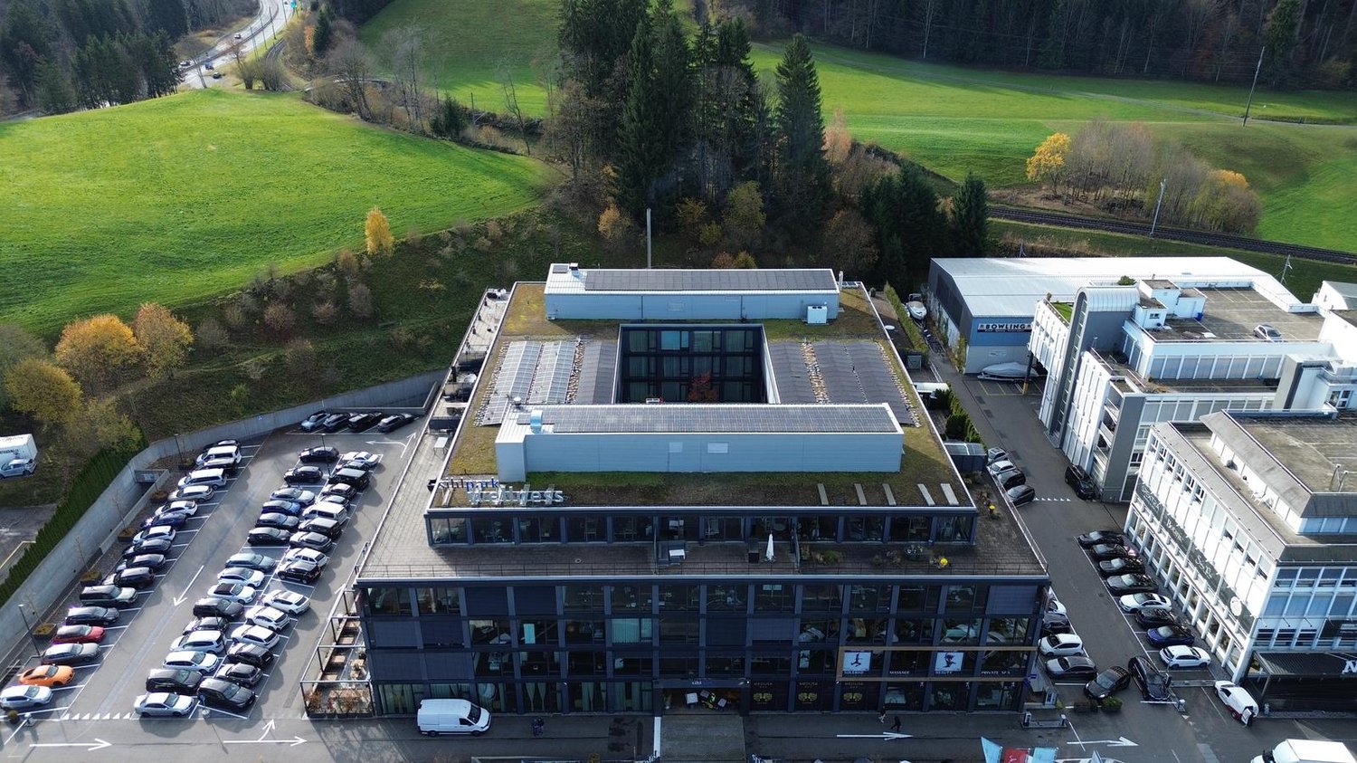 large building, solar panels on the roof, lots of cars parked, green areas, surrounded by a fence