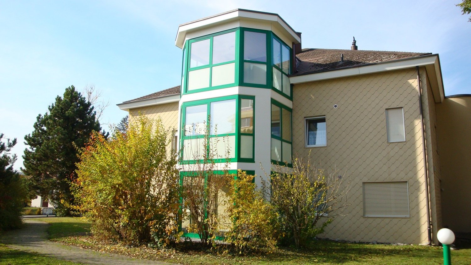 Two story house, green and beige exterior, large bay windows, trees and shrubs in front