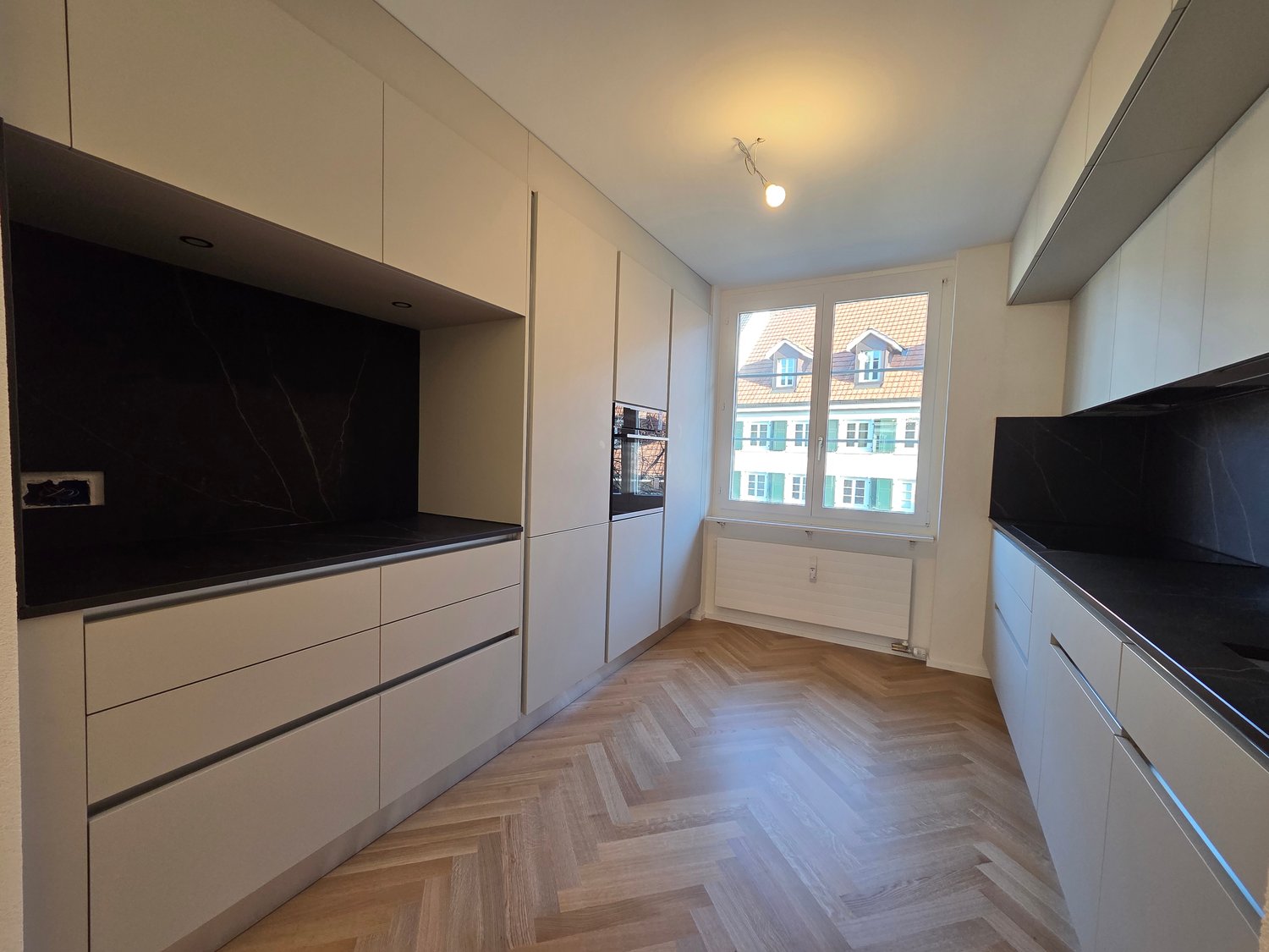 Modern kitchen with white cabinetry, wooden herringbone flooring, stainless steel countertop, large window with view of the city, ceiling light, integrated oven, white floor radiator, and white cabinetry