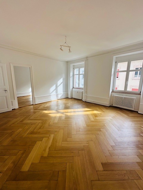 An empty room with parquet flooring, white walls, three windows, and two radiators. One of the rooms has a mirror.