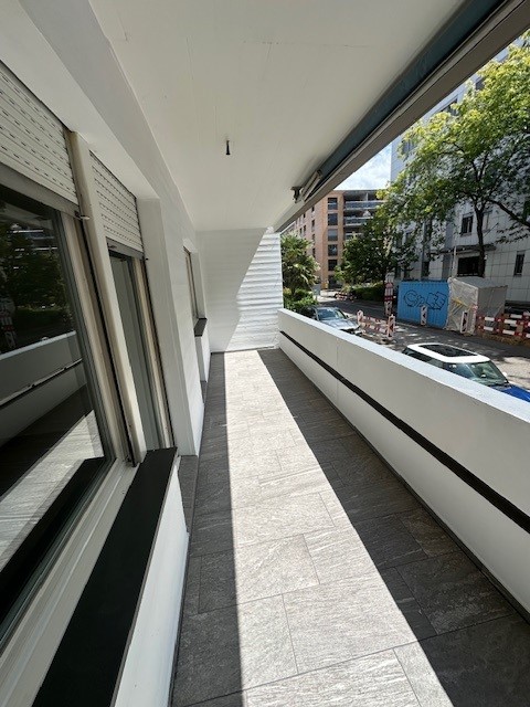 Apartment building balcony, white railings, tiled floor, shadow on the ground, windows on the left side, and buildings and cars on the road on the right side.