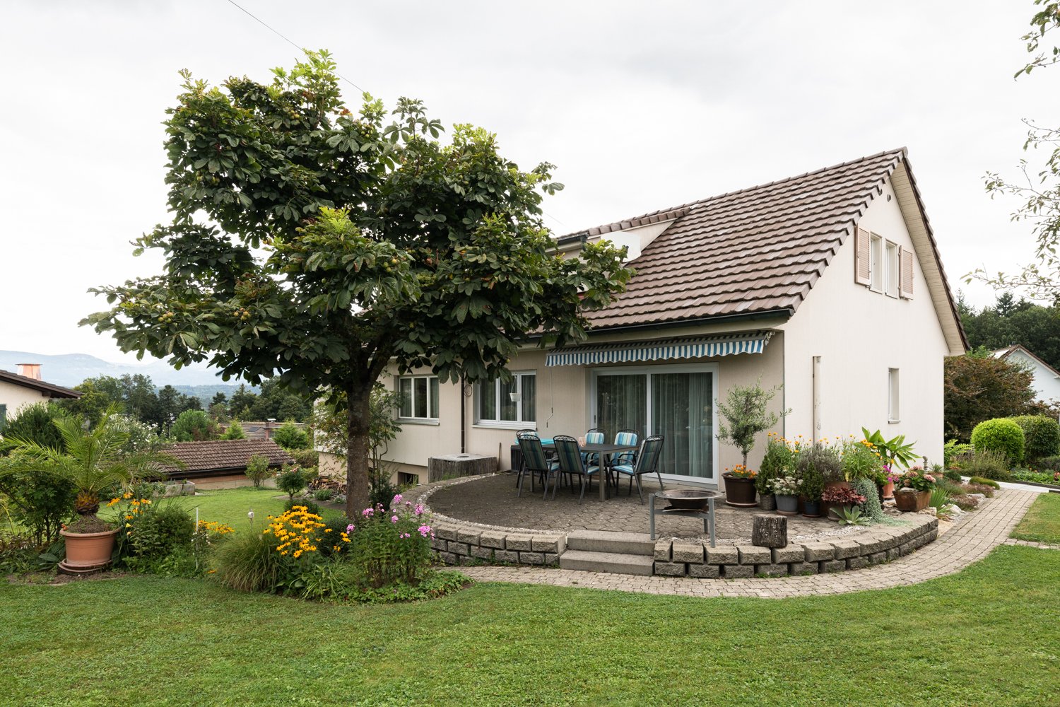 Two-story house with a tiled roof, surrounded by a well-maintained garden with a paved patio area and outdoor seating. The house has large windows and a covered balcony or terrace. There is also a parking area near the house.