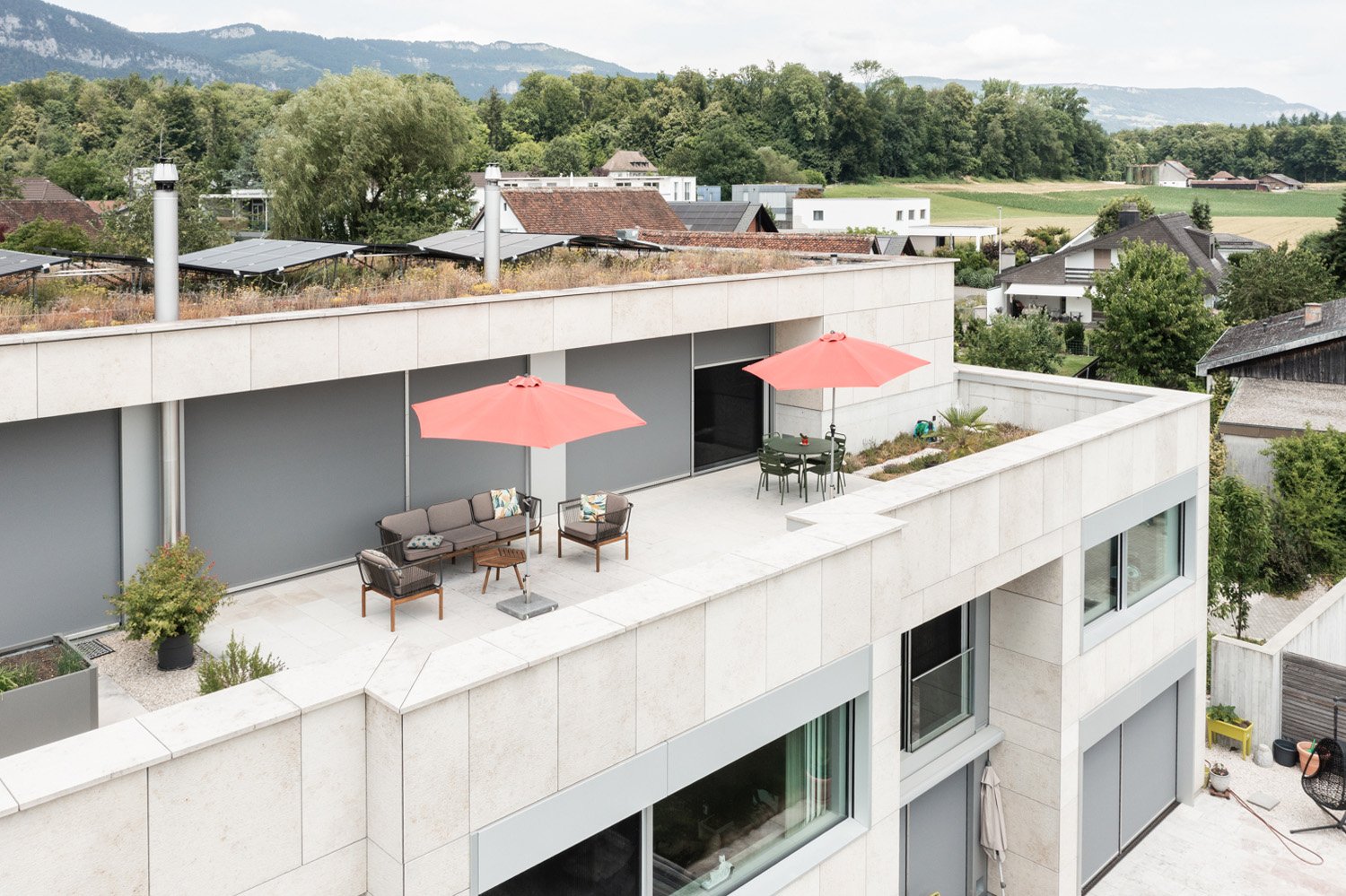 Modern white building with green roof terrace, solar panels, outdoor furniture, and an umbrella
