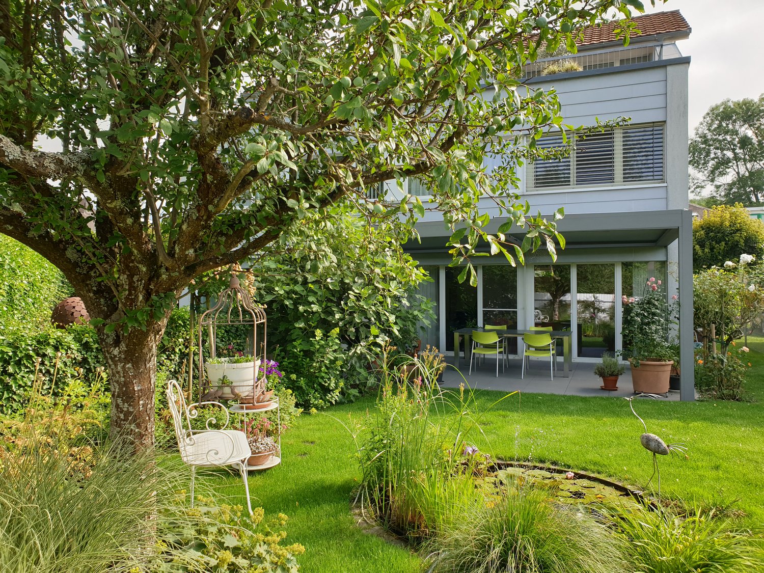 garden with a green lawn, patio with table and chairs, potted plants, bird statue, small pond, and various plants