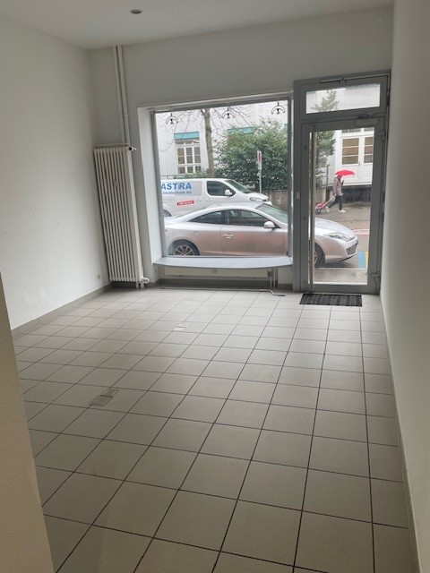 The image shows an empty commercial space with a tiled floor and large windows. Through the windows, a parked car can be seen, indicating a parking place or garage nearby.