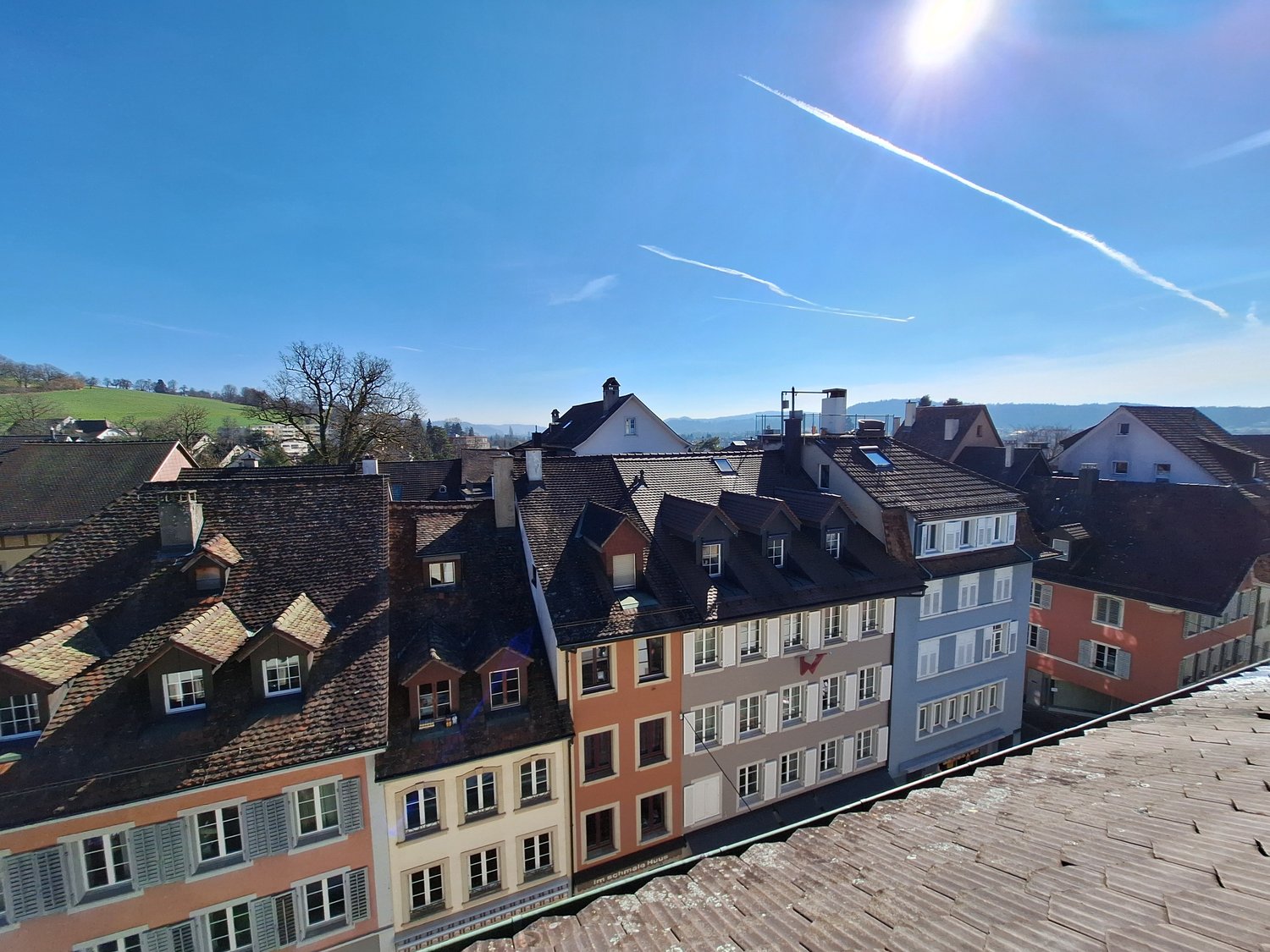 The image shows a cityscape with a view of the rooftops of old, colorful buildings in a European style. The buildings have a mix of architectural styles, including pitched roofs and dormers. The sky is clear and blue, with contrails visible overhead. The