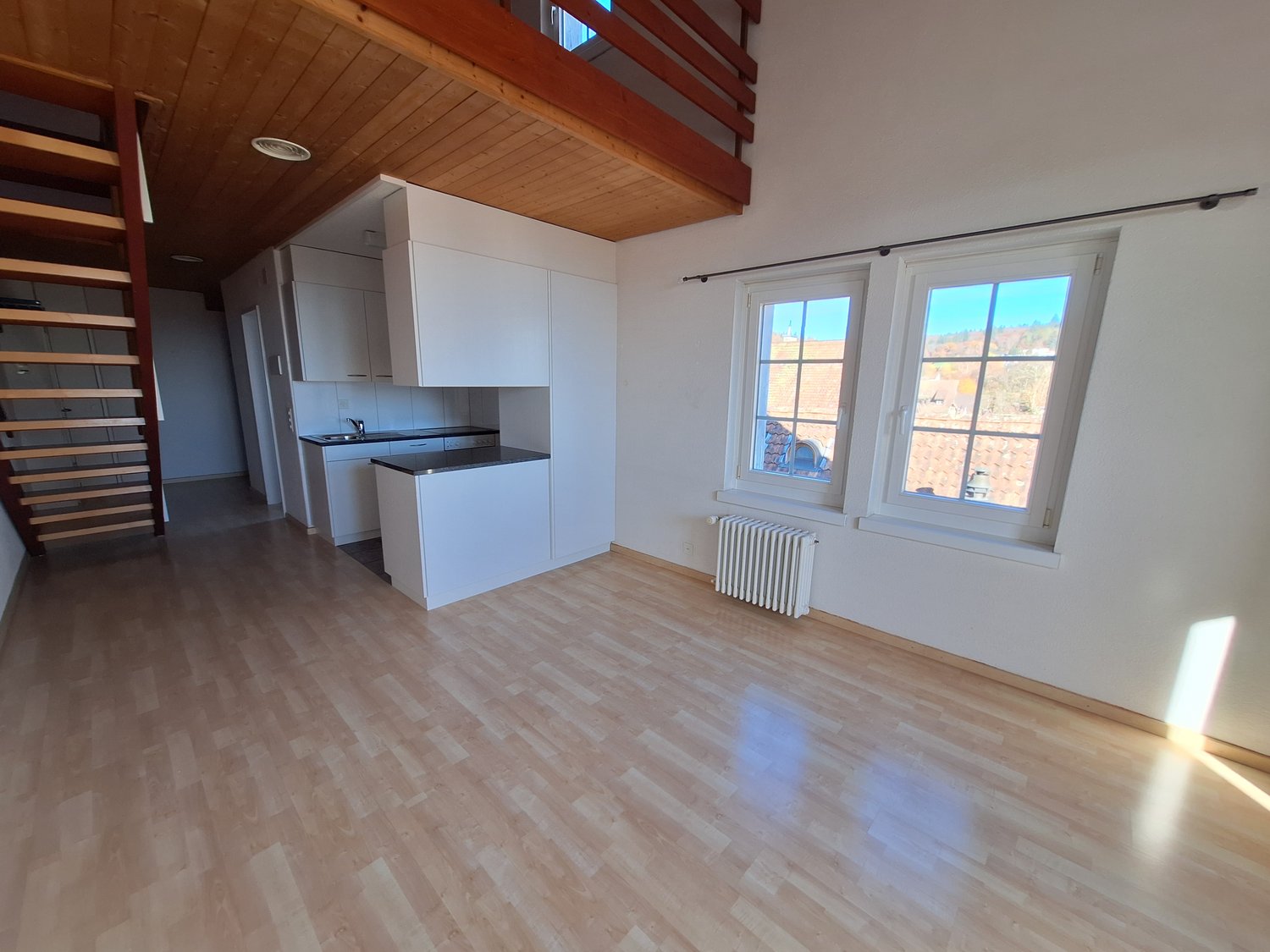 Empty room, wooden floor, white walls, wooden ceiling, wooden staircase, open kitchen, two windows
