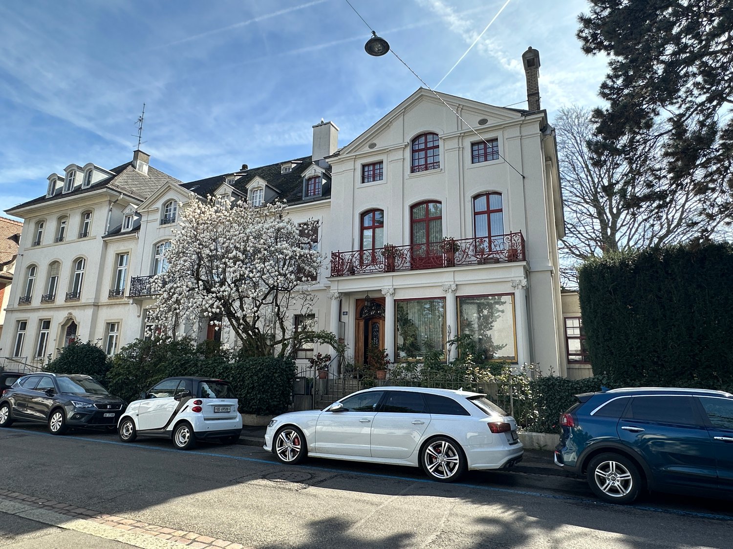 A large, multi-story building with a classical architectural style, featuring arched windows, a balcony, and a chimney. The building is surrounded by trees and parked cars in the foreground.