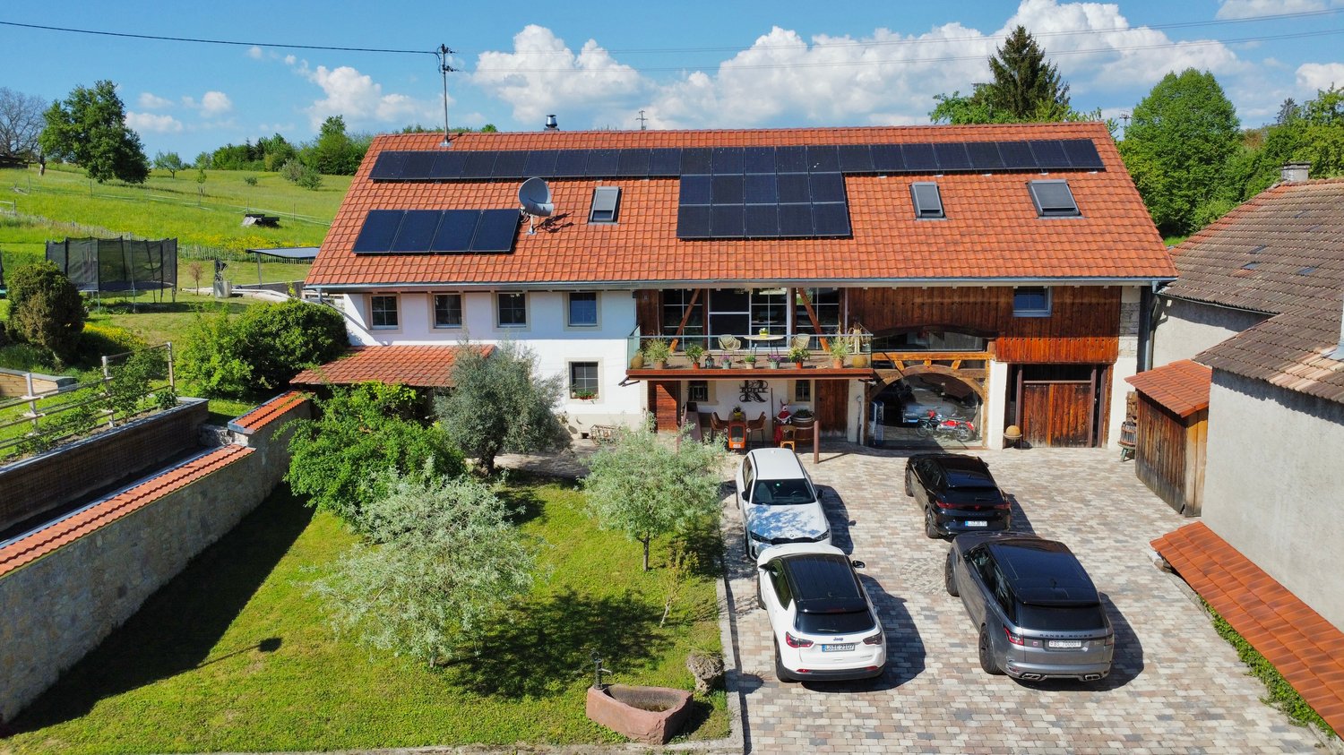2-story house with red tile roof, solar panels installed on the roof, balcony, garage, and parking spaces in the courtyard, surrounded by greenery and trees