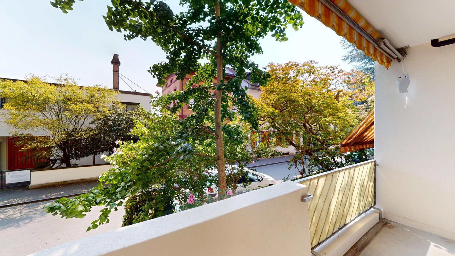 A balcony with white railings and a yellow awning, overlooking a street with trees, flowers, and buildings in the distance. A car is parked on the street.