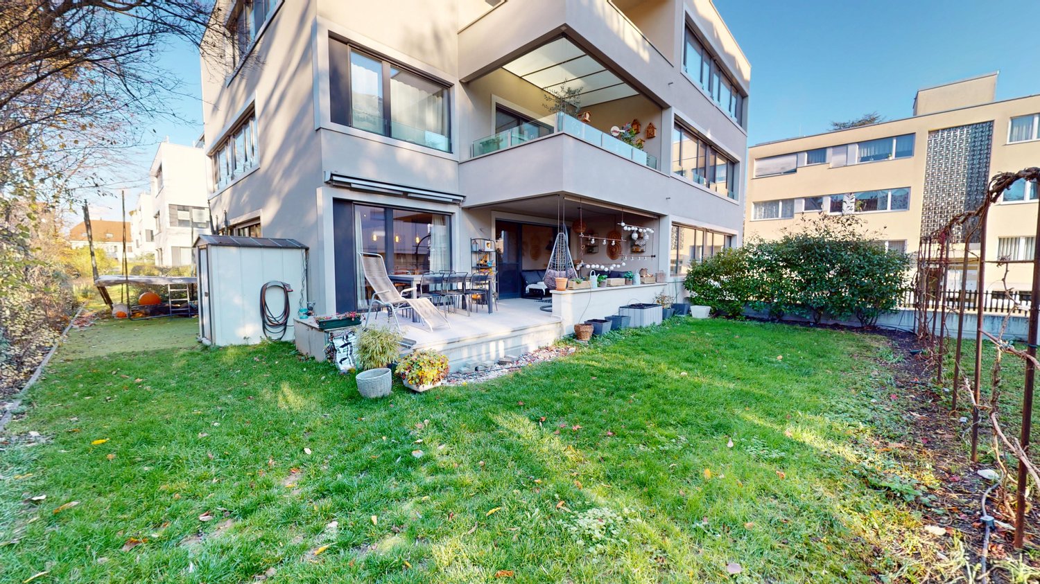 modern apartment building, well-maintained garden, wooden chairs on porch, glass balcony, neatly trimmed grass