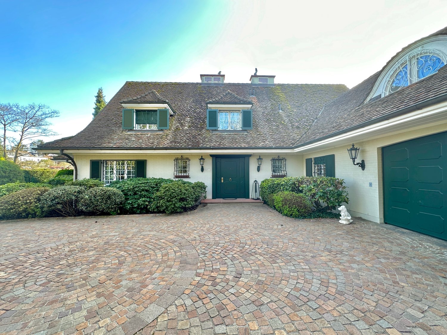 traditional style house, green front doors, brick driveway, green garage doors, lamps, bushes, statue, trees in the background