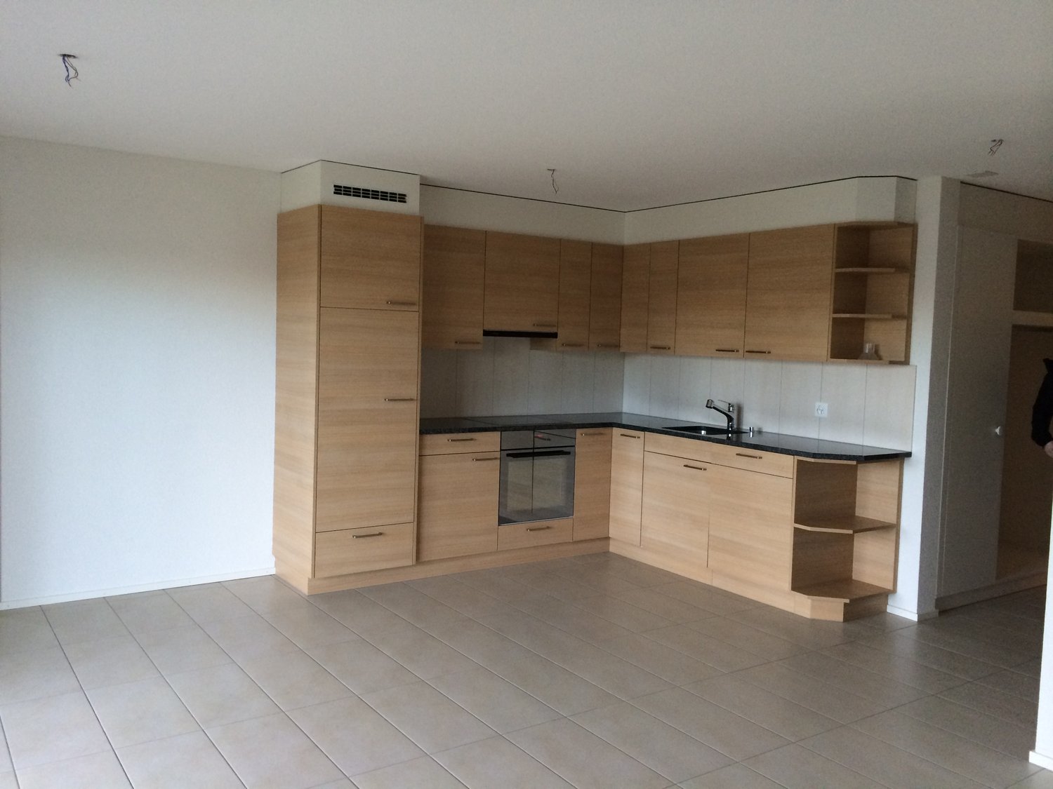 Spacious L-shaped kitchen with light wood cabinets, built-in oven, and dishwasher. The kitchen has a clean, modern design with a tiled floor and white walls.