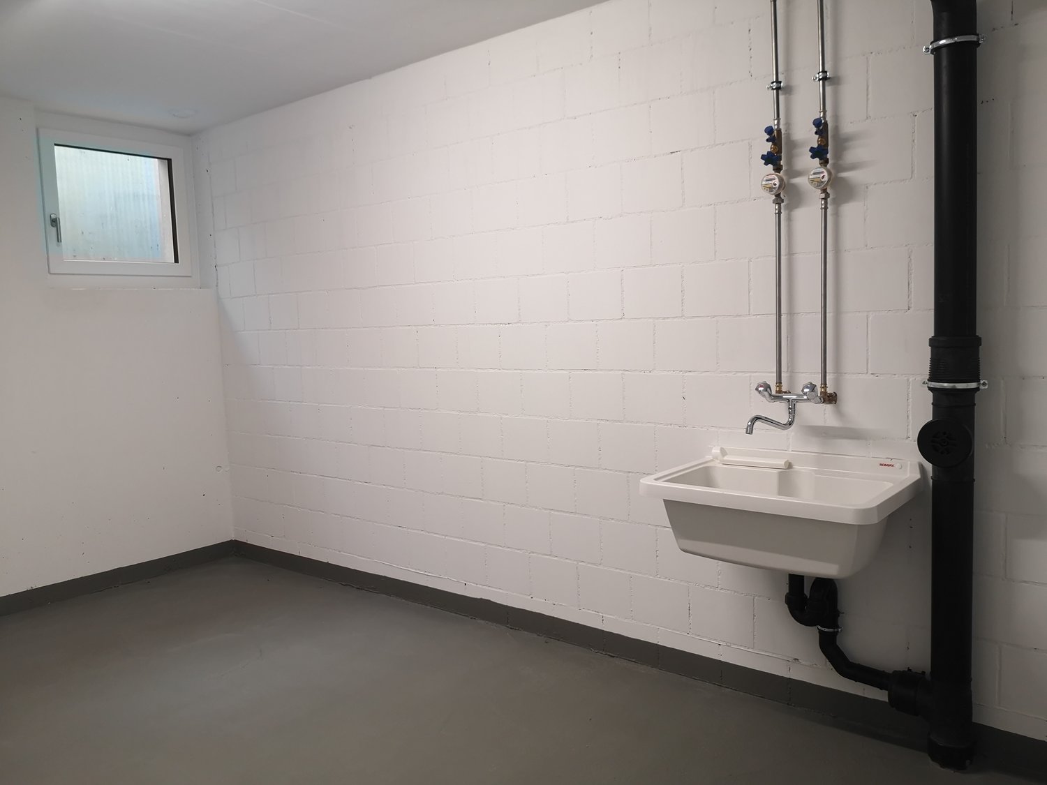 An empty basement with white brick walls, concrete floor, small sink, and pipes.